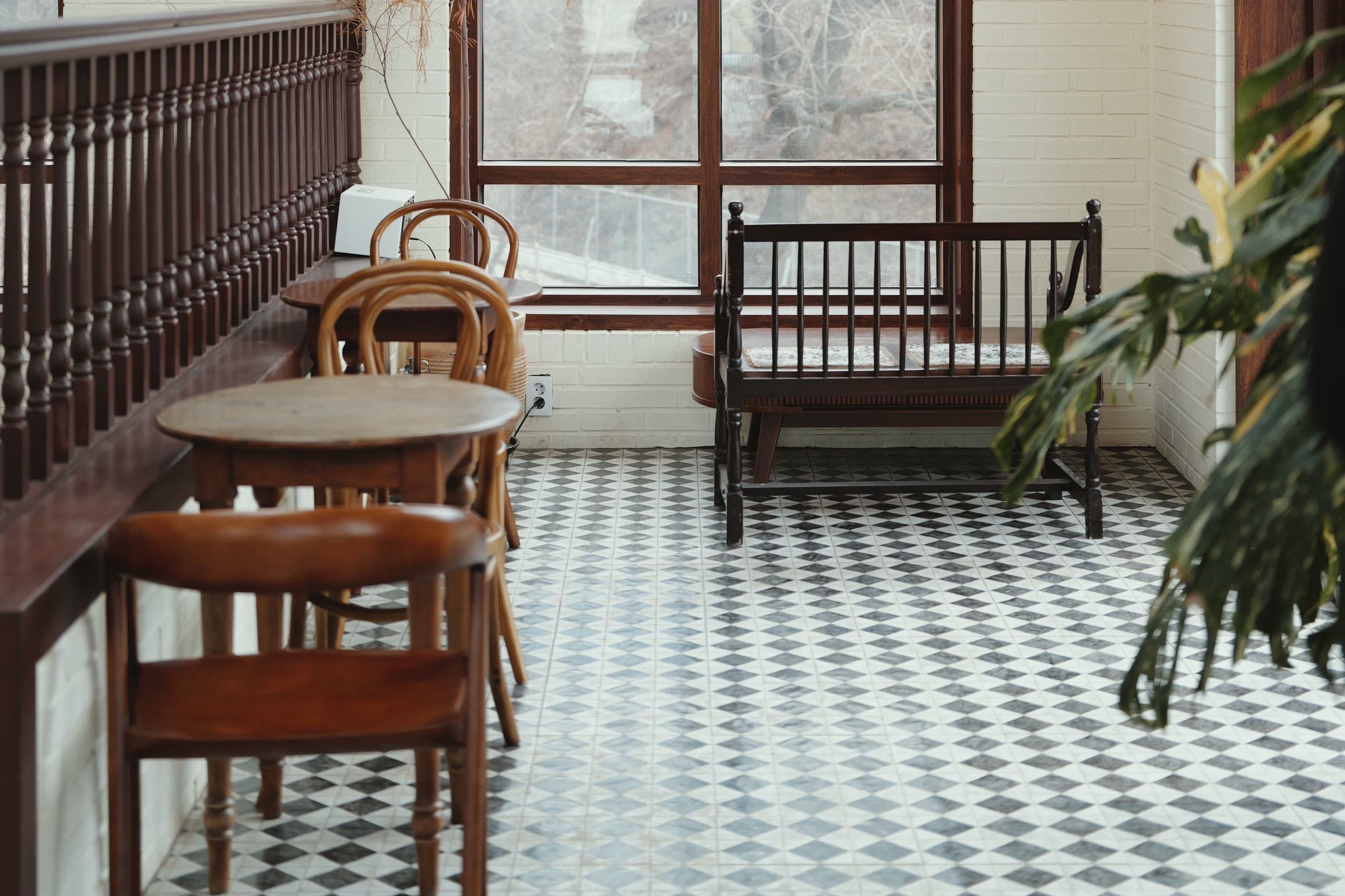 A sunlit café corner with a black-and-white checkered tile floor, round wooden tables, bentwood chairs, and an antique wooden bench