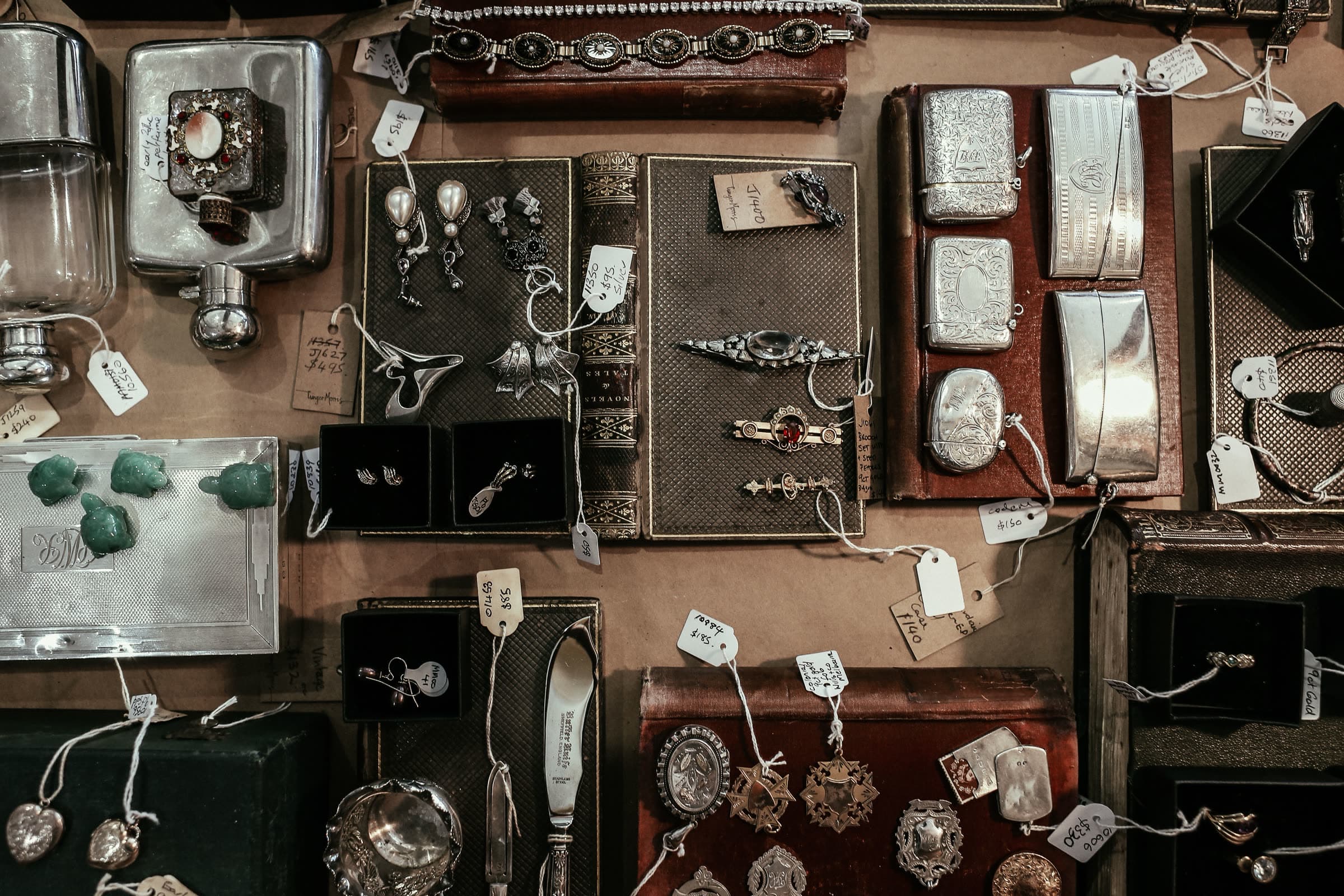 Antique silver collectibles — brooches, earrings, engraved cases, and flatware — laid out on old book covers with handwritten price tags