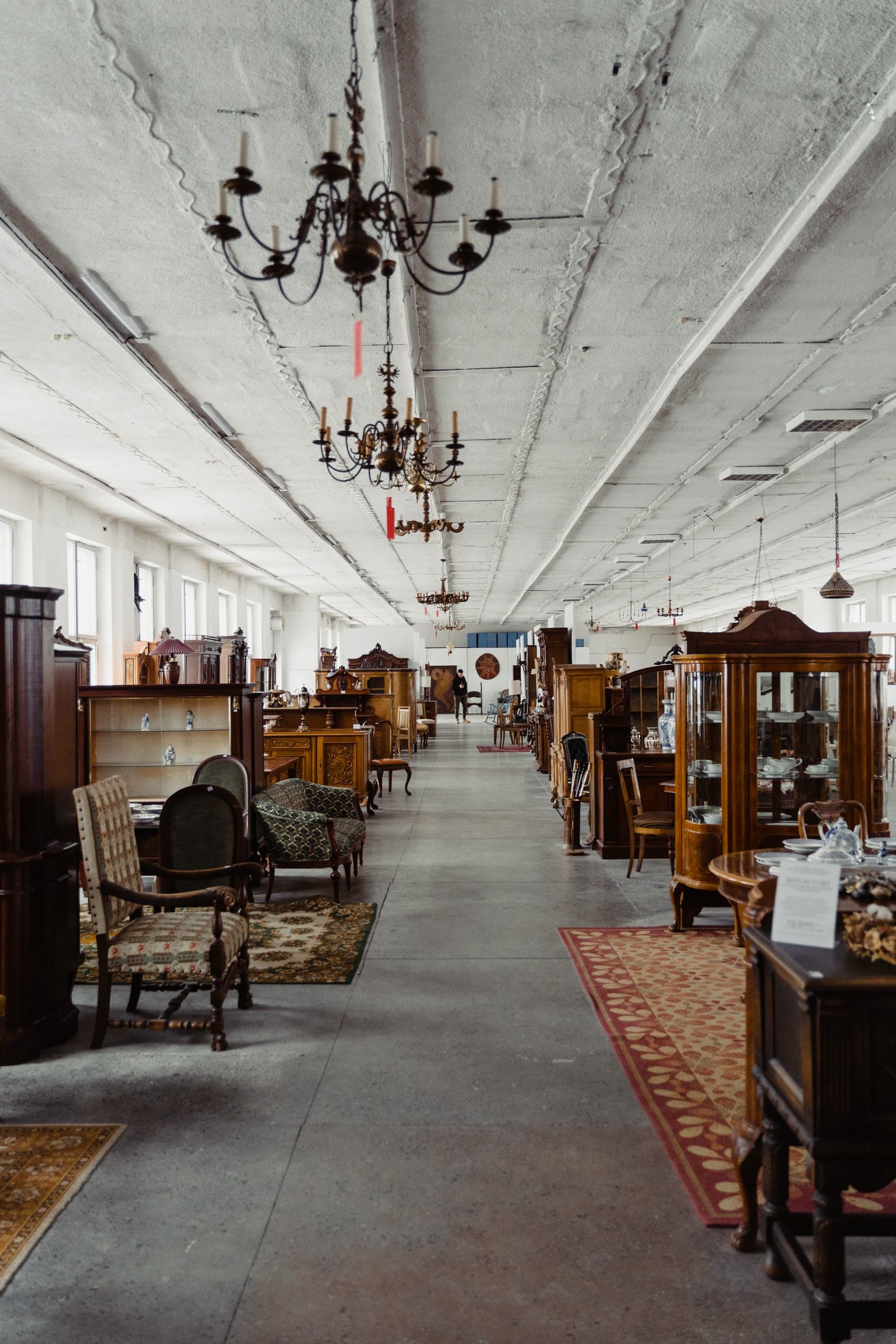 A long aisle of antique furniture — carved cabinets, upholstered armchairs, and glass-front display cases — under hanging chandeliers